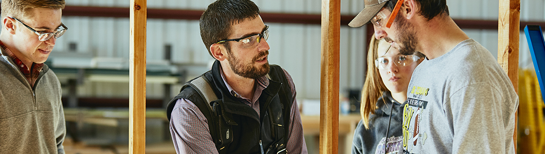 A group of students in protective equipment discuss coursework in a barn.