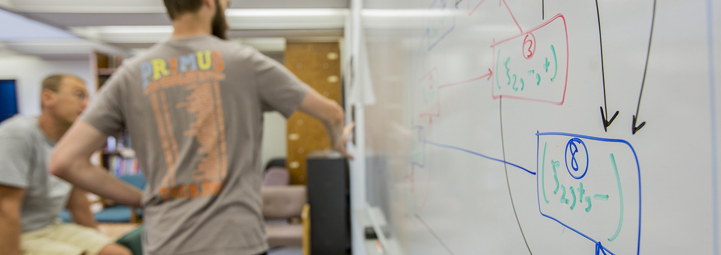 A young man in a ball cap and braided pigtails is in deep discussion with another classmate. Equations are seen in the background on the whiteboard.