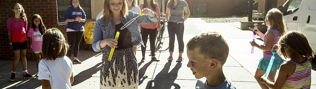 A teacher blows bubbles for a group of children on a playground.