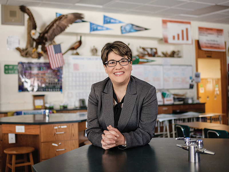 A person wearing a gray blazer and glasses with short brown hair smiling to the camera