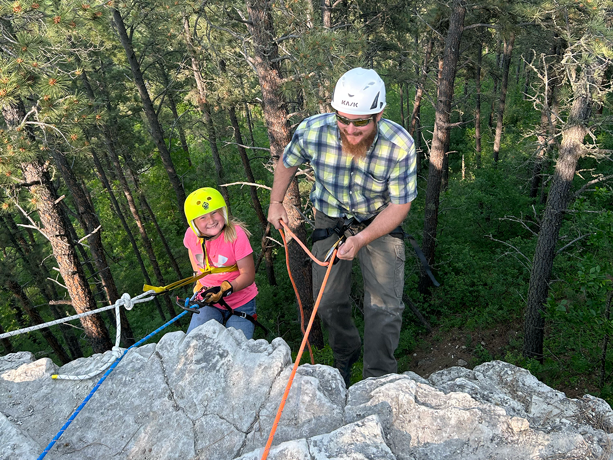 A young girl wearing a pink shirt and neon yellow helmet next to a man with a plaid shirt white helmet and sunglasses, holding ropes as they go down a small cliffside