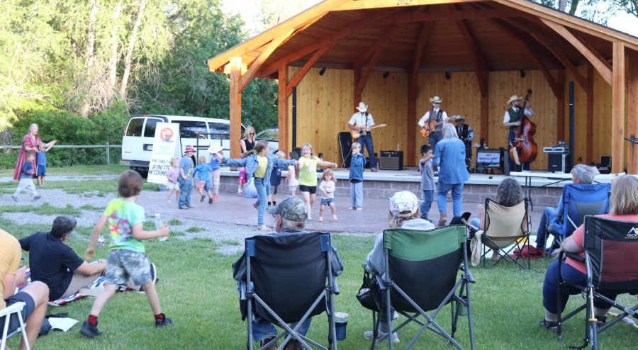 Spectators in camp chairs watch and children dance in front of a wooden bandstand with a 4-person band playing inside