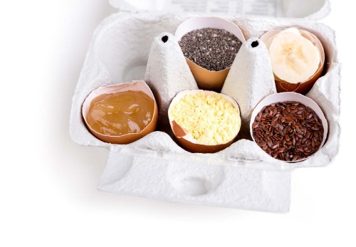 A carton for six eggs holds 5 half shells. Each half contains a different egg substitute, such as chia seeds, flax seeds, banana, applesauce, and egg replacer powder.