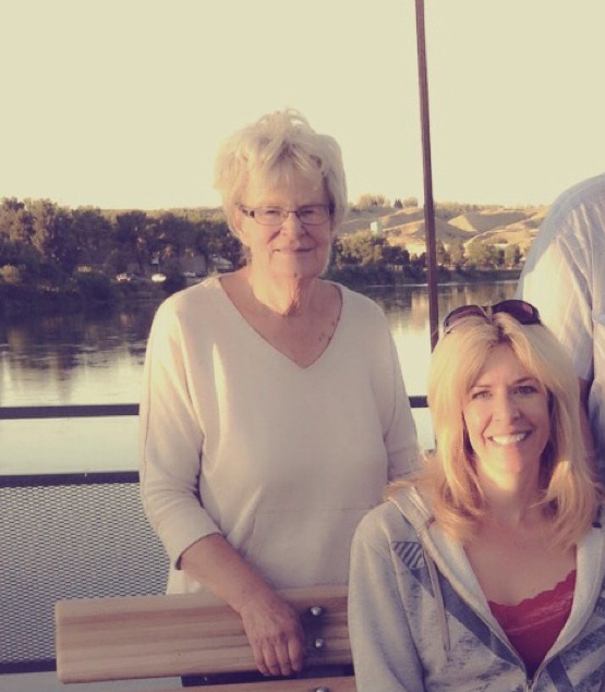 Darla, an older woman, stands behind another woman sitting on a bench.