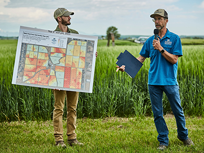 Two people stand in a grassy field during an outdoor agricultural event. One person holds a large map showing color-coded field plots, while the other speaks into a microphone and holds a folder. Tall green crops and a cloudy sky are visible in the background.