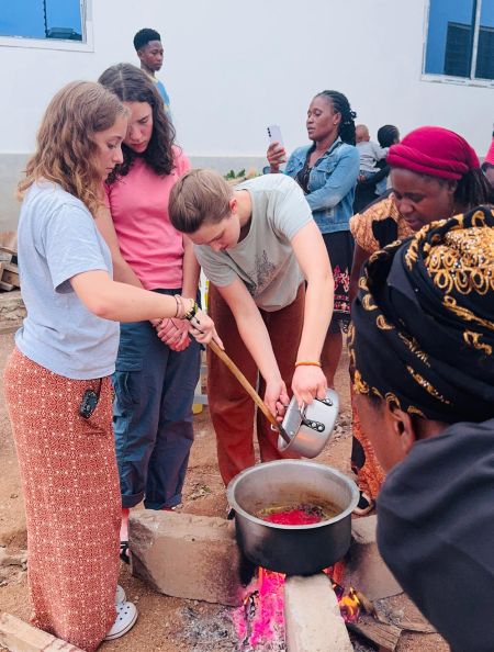 Several people gathered around an outdoor cooking fire, with one person stirring a pot and another pouring ingredients into it.