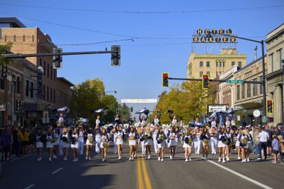Cheerleaders perform in a homecoming parade on a city street lined with spectators and historic buildings.