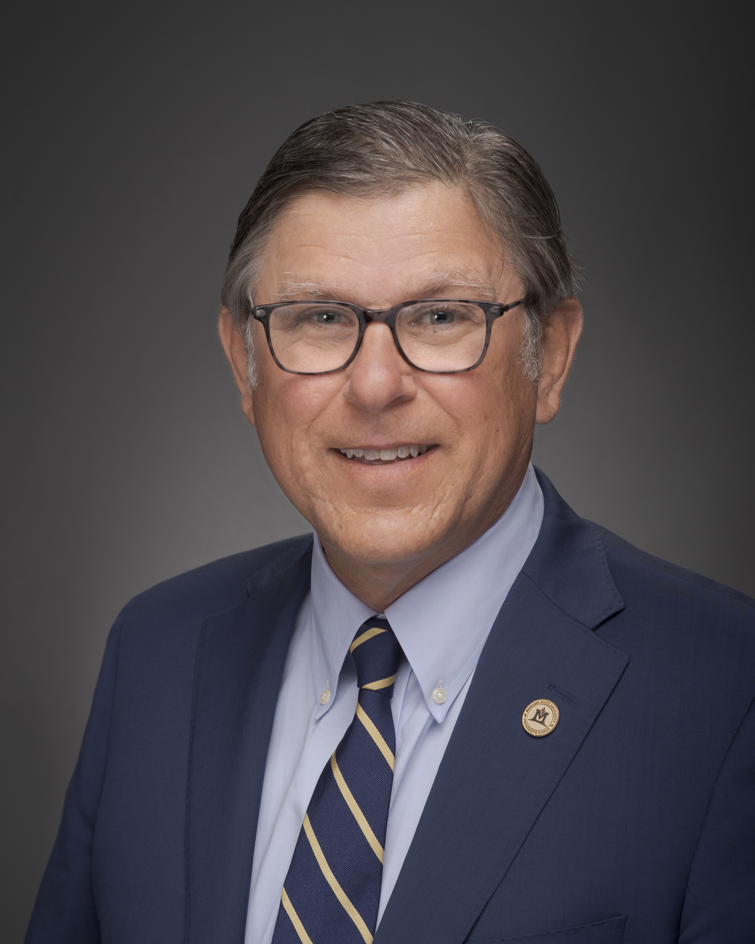 The image shows a man with short, dark hair and glasses, smiling while looking at the camera. He is wearing a dark blazer over a white shirt with a blue checkered pattern. The background is black.
