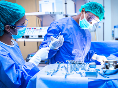 A surgical technologist stands over a tray of surgical tools as she hands a surgeon one of the implements while a patient is being operated on.