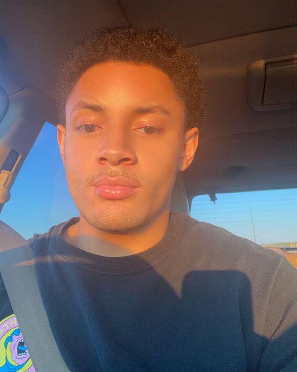 A person with short black hair wearing a black shirt with a seatbelt across their chest