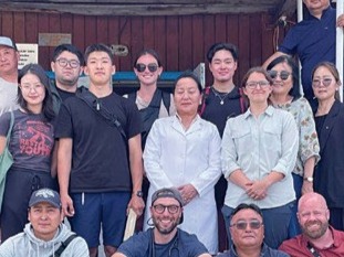 A group of women and men standing in front of a wooden building, looking straight ahead. 