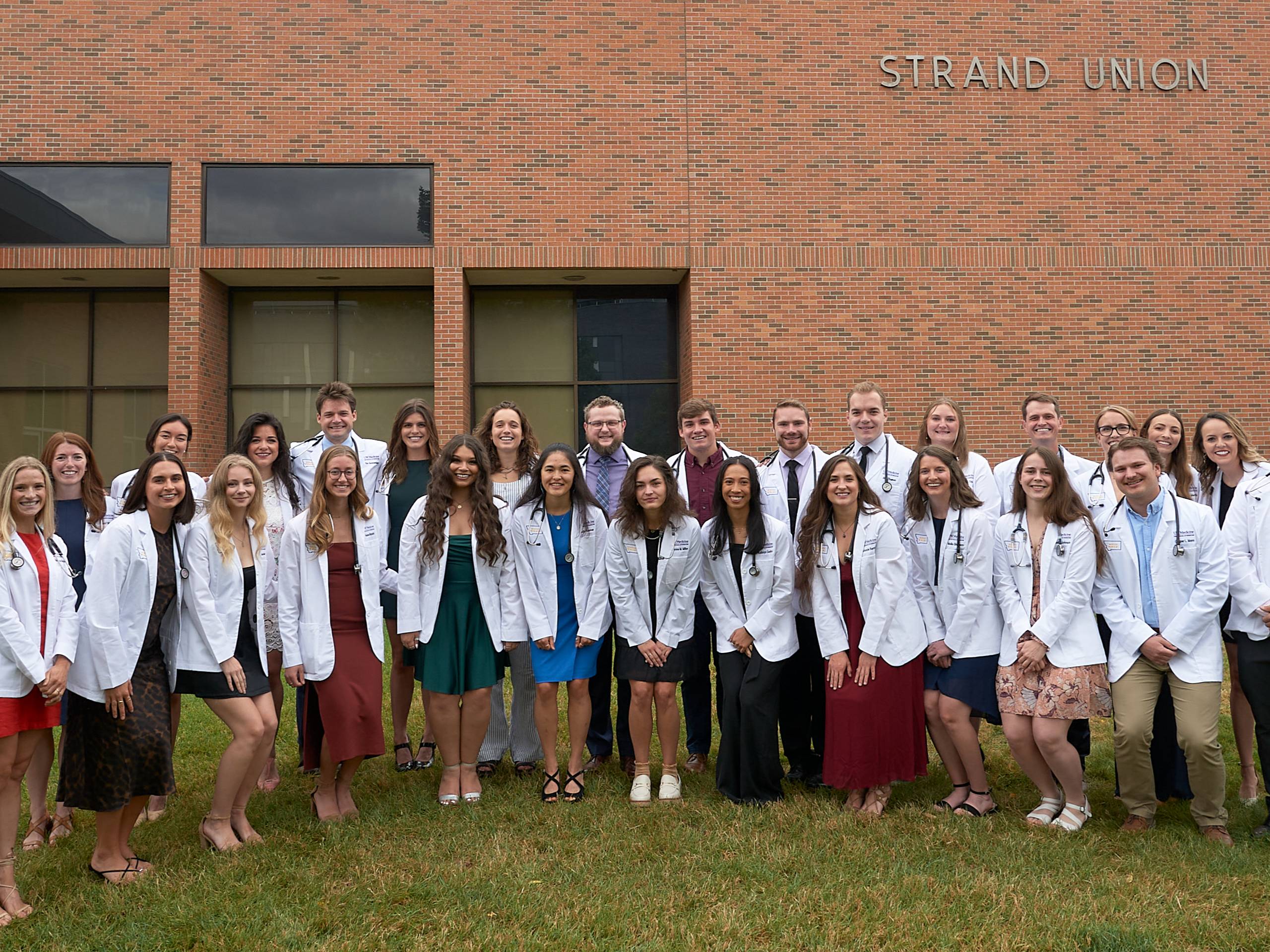 A group of young men and women wearing white lab coats, posing on the grass in front of a brick building, smiling and looking straight ahead. 